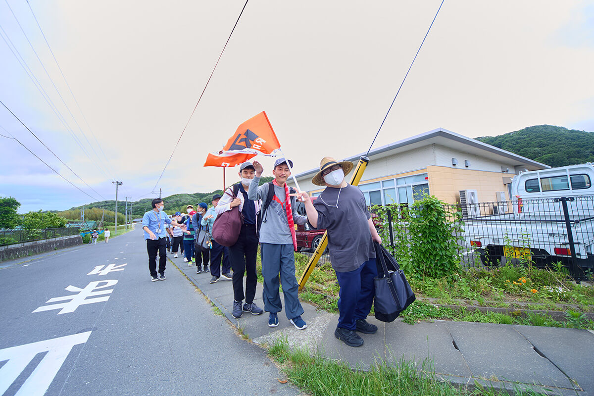 グループホームへの帰り道、オレンジ色の旗を掲げ、施設前の道を歩く利用者たちの写真