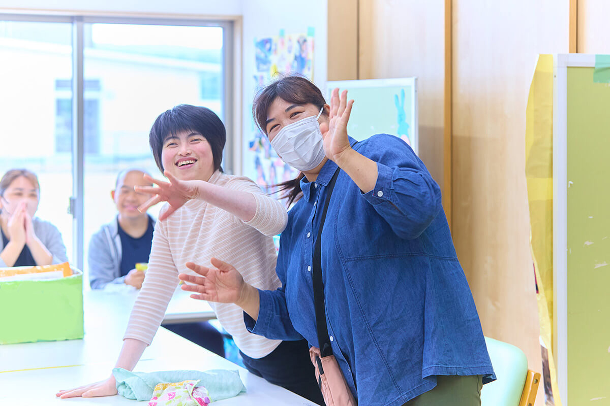 室内で両手を広げ、笑顔でポーズをとる利用者と職員の写真